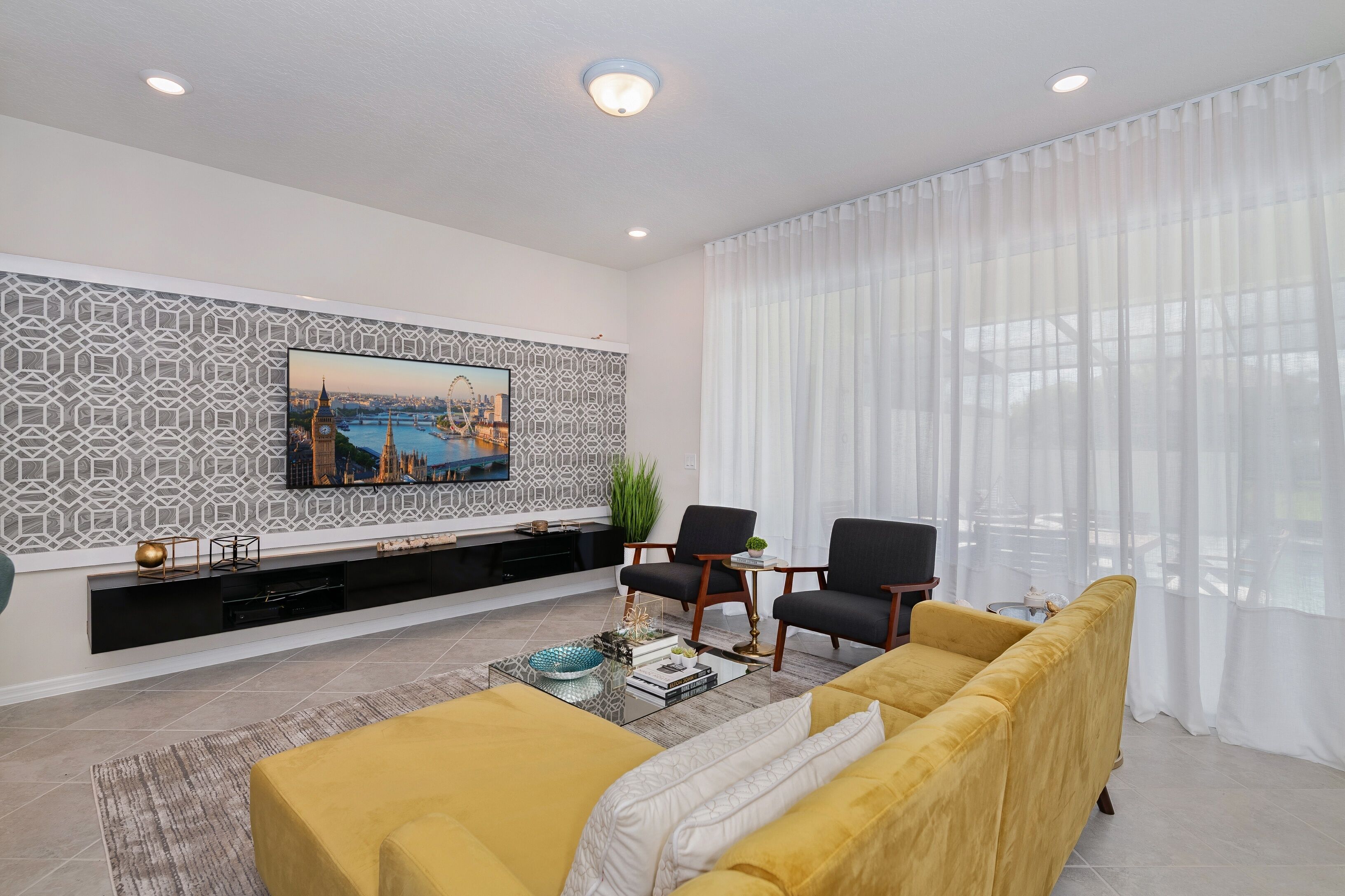 A modern living room with a large flat-screen TV mounted on a patterned wall, a yellow sofa, two black armchairs, a coffee table with books and decorative items, and a large window with sheer white curtains.
