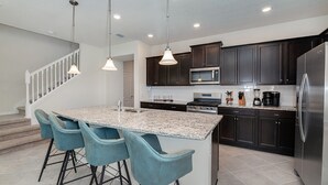 A modern kitchen with dark cabinets, a stainless steel refrigerator, a microwave, and a granite island with bar stools. - Sun Traveler @ Solterra By Shine Villas #113 5 Bedroom Townhouse (Davenport)