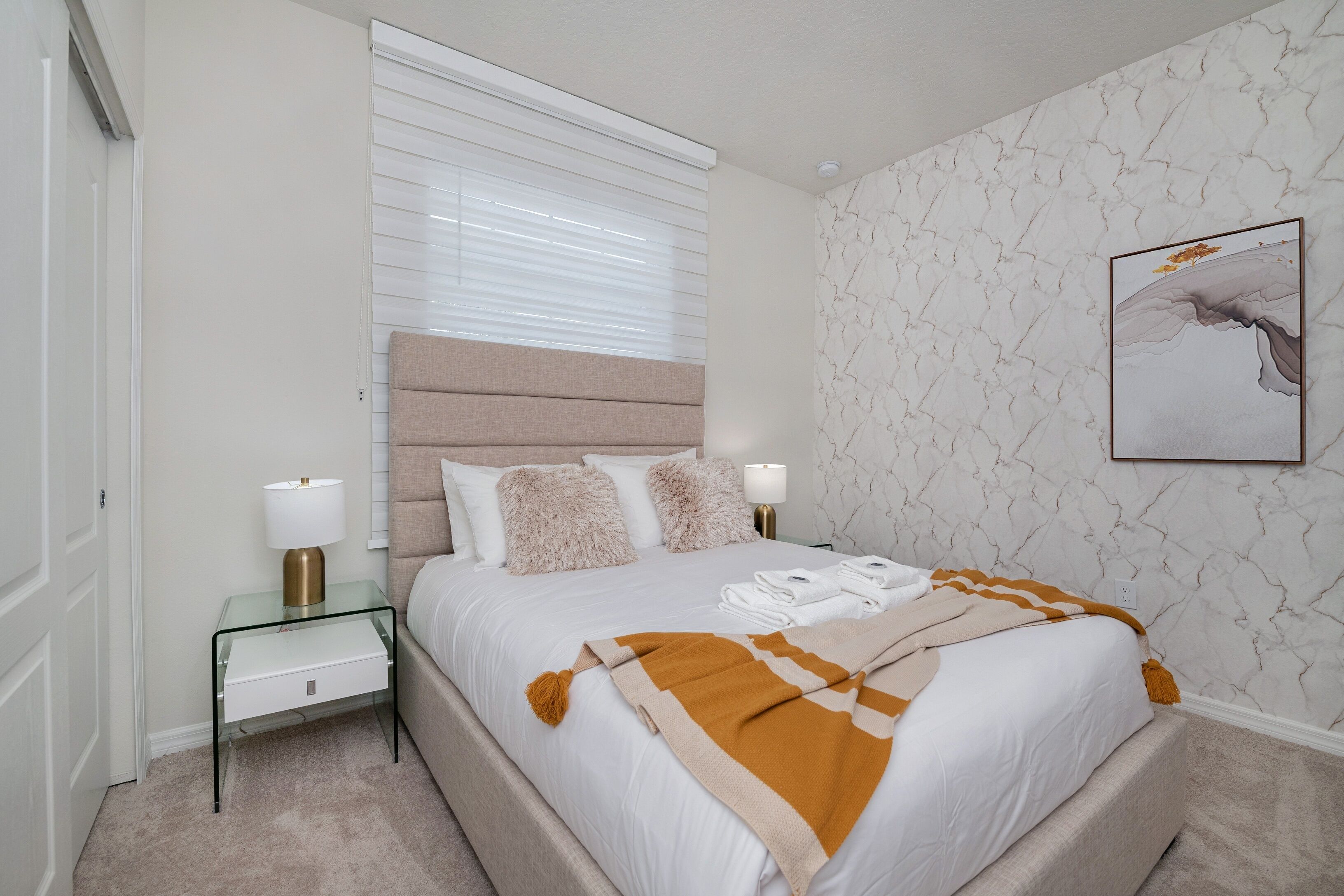 Een netjes ingerichte slaapkamer met een bed, nachtkastjes, een raam met zonwering, een wand met marmerpatroon en een ingelijste foto.