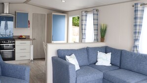 A modern living room with a blue sofa, a kitchen area with a stove, and a window with curtains. - The Retreat (Budleigh Salterton)