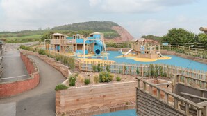A playground with wooden structures and slides, surrounded by a brick wall and a wooden fence. - The Retreat (Budleigh Salterton)