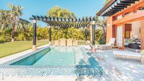 Zona de piscina con una pérgola, mobiliario exterior y un edificio moderno con grandes ventanales.