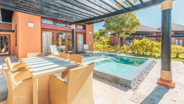 Un área junto a la piscina con una pérgola, mobiliario de mimbre y una piscina cristalina.