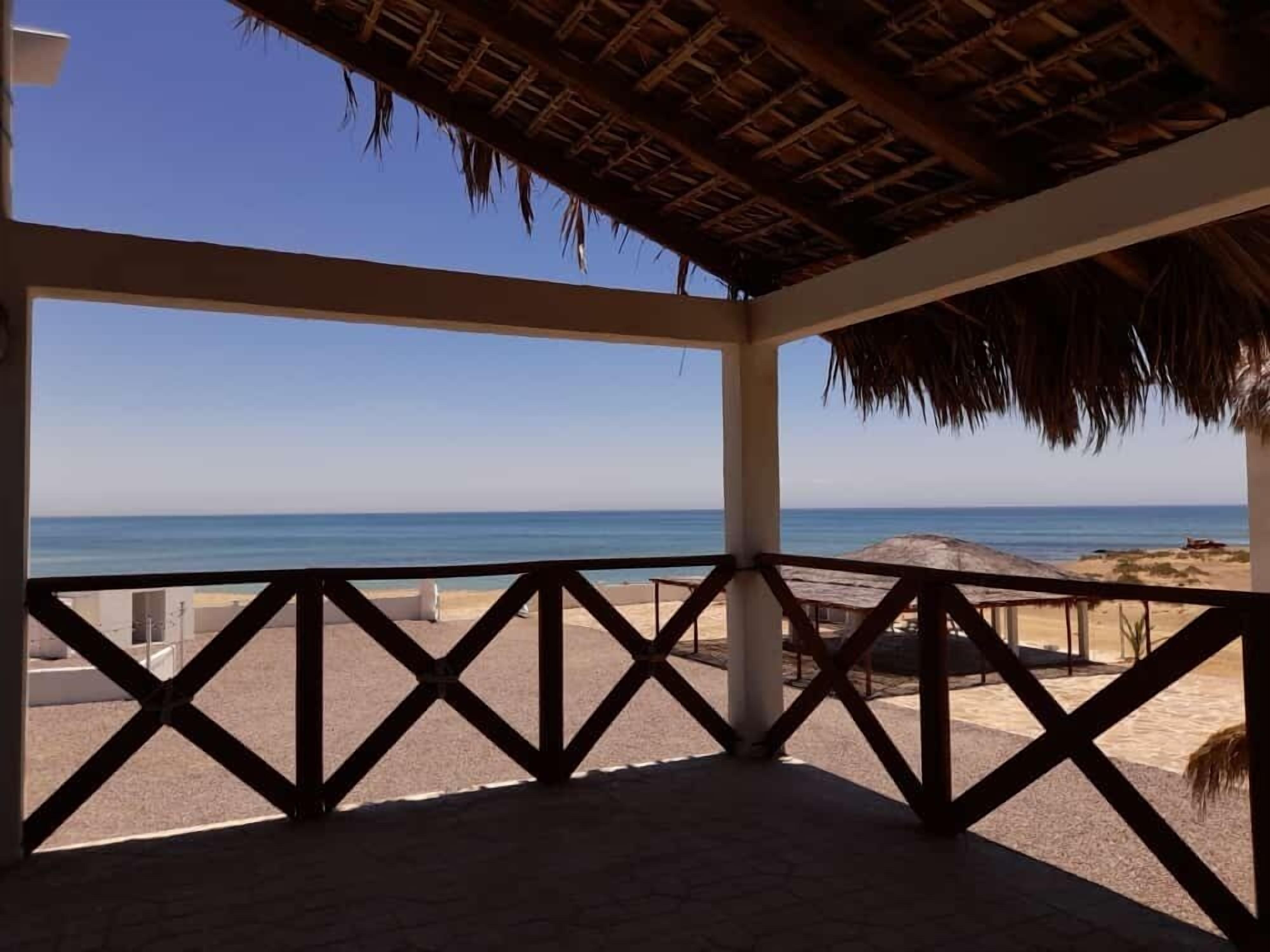A beach view from a covered area with a thatched roof, featuring a clear blue sky and calm sea.