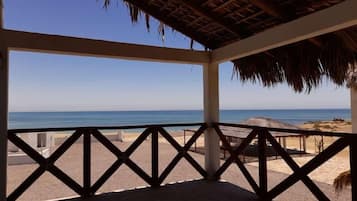 A beach view from a covered area with a thatched roof, featuring a clear blue sky and calm sea.