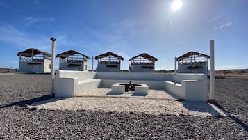 A row of white cabins with thatched roofs, arranged in a line on a gravel surface, under a clear blue sky.