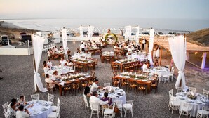 Une réception de mariage en plein air, avec des tables rondes et des chaises blanches, aménagée sur une surface de gravier offrant une vue sur l’océan.