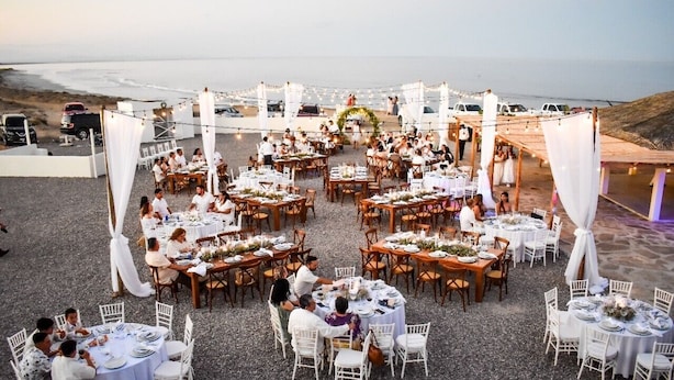 Une réception de mariage en plein air, avec des tables rondes et des chaises blanches, aménagée sur une surface de gravier offrant une vue sur l’océan.