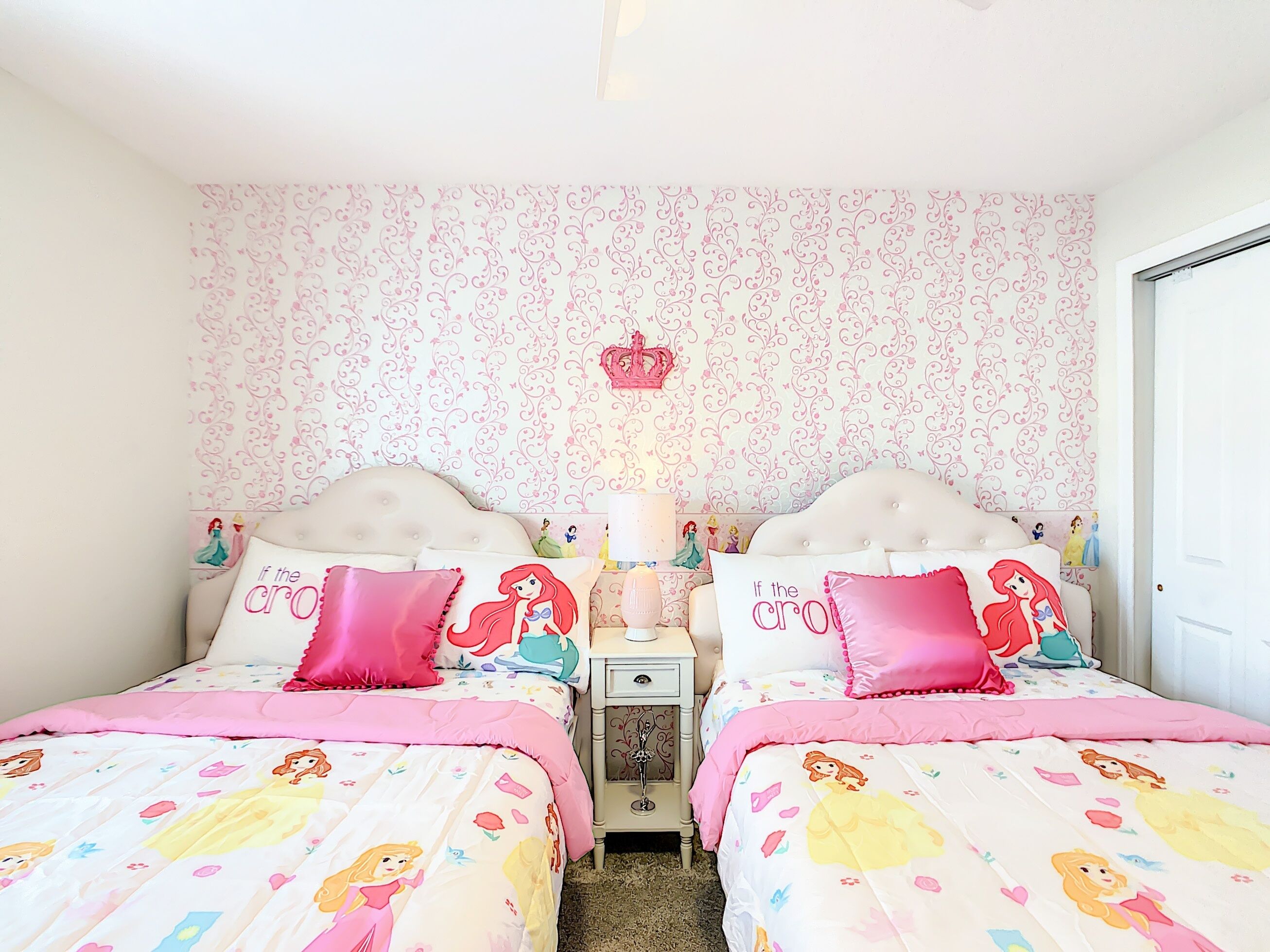 Une chambre avec deux lits, chacun recouvert d’une couette à motifs de princesses, une table de chevet avec une lampe et un mur orné d’un motif floral et d’une décoration en forme de couronne.