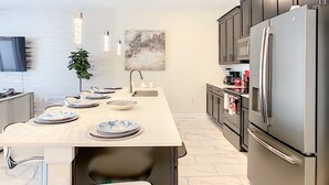 A modern kitchen with a central island, stainless steel appliances, pendant lighting, and a flat-screen TV.