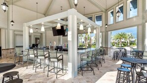 A spacious outdoor bar area with multiple seating arrangements, including tables and bar stools, under a high ceiling with hanging lights.