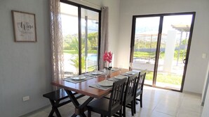 Uma área de refeições com uma mesa de madeira posta para quatro pessoas, um banco e vista para uma piscina através de grandes janelas.