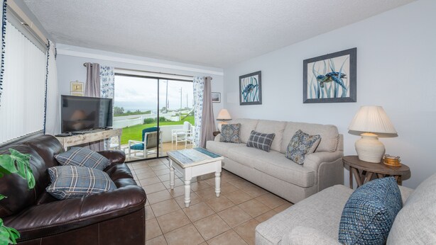 A living room with a sofa, a coffee table, and a TV. There is a view of the outdoors through a glass door. - Maria Beautiful Beachfront Condo, sleeps up to 8. BIG DISCOUNTS NOW  (Flagler Beach)