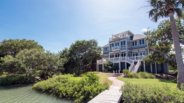 A multi-story house with a dock leading to a body of water, surrounded by lush greenery and palm trees.