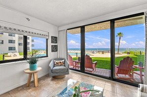 A balcony with a view of a beach and pool, a grey armchair, a small round table with a plant, and a large window.