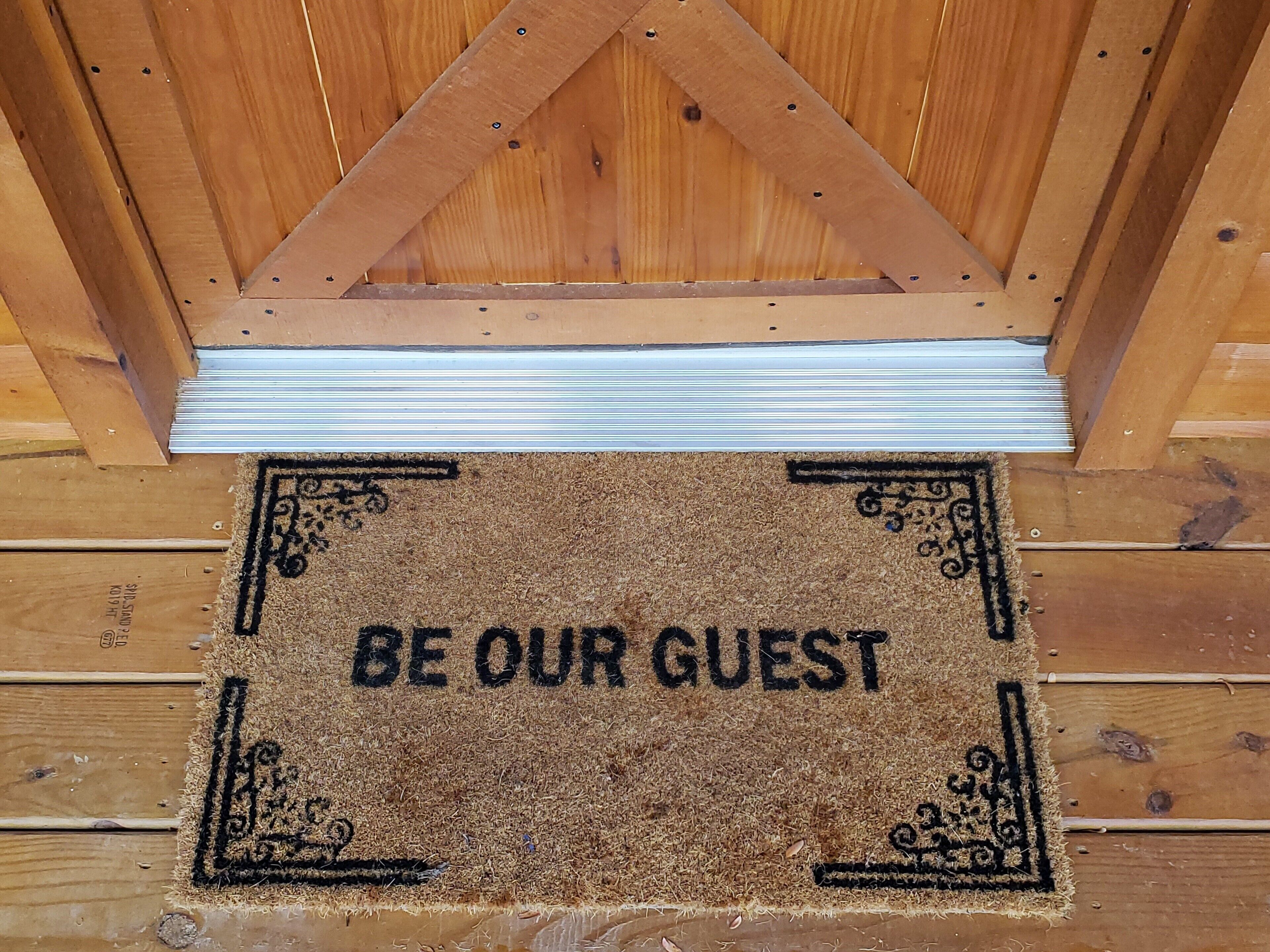 Una puerta de madera con un felpudo que dice “BE OUR GUEST”.