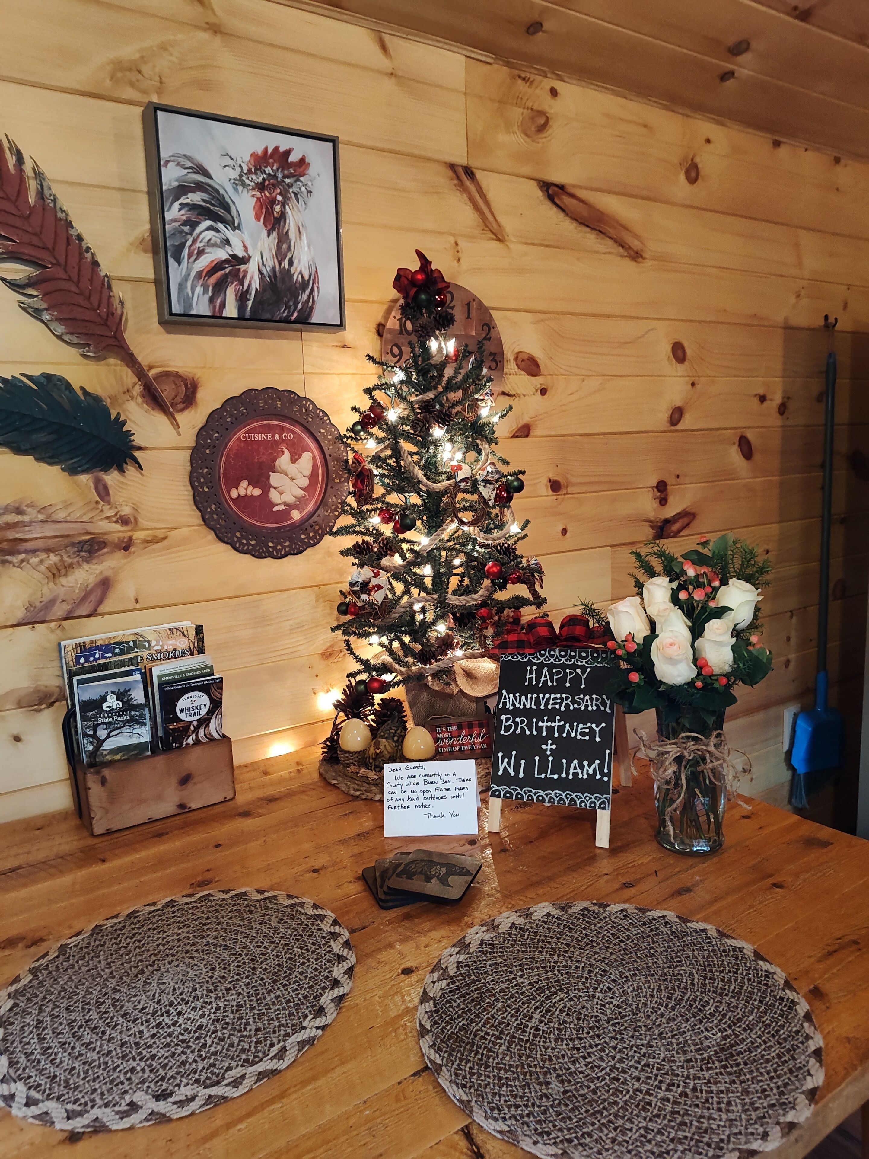 Een houten interieur met een versierde kerstboom, een kunstwerk van een haan in een lijst, een klok en een bordje voor het jubileum van Brittney & William.