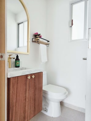 A bathroom with a white toilet, a wooden vanity with a marble countertop, a gold-framed mirror, and a small shelf with a vase of flowers.