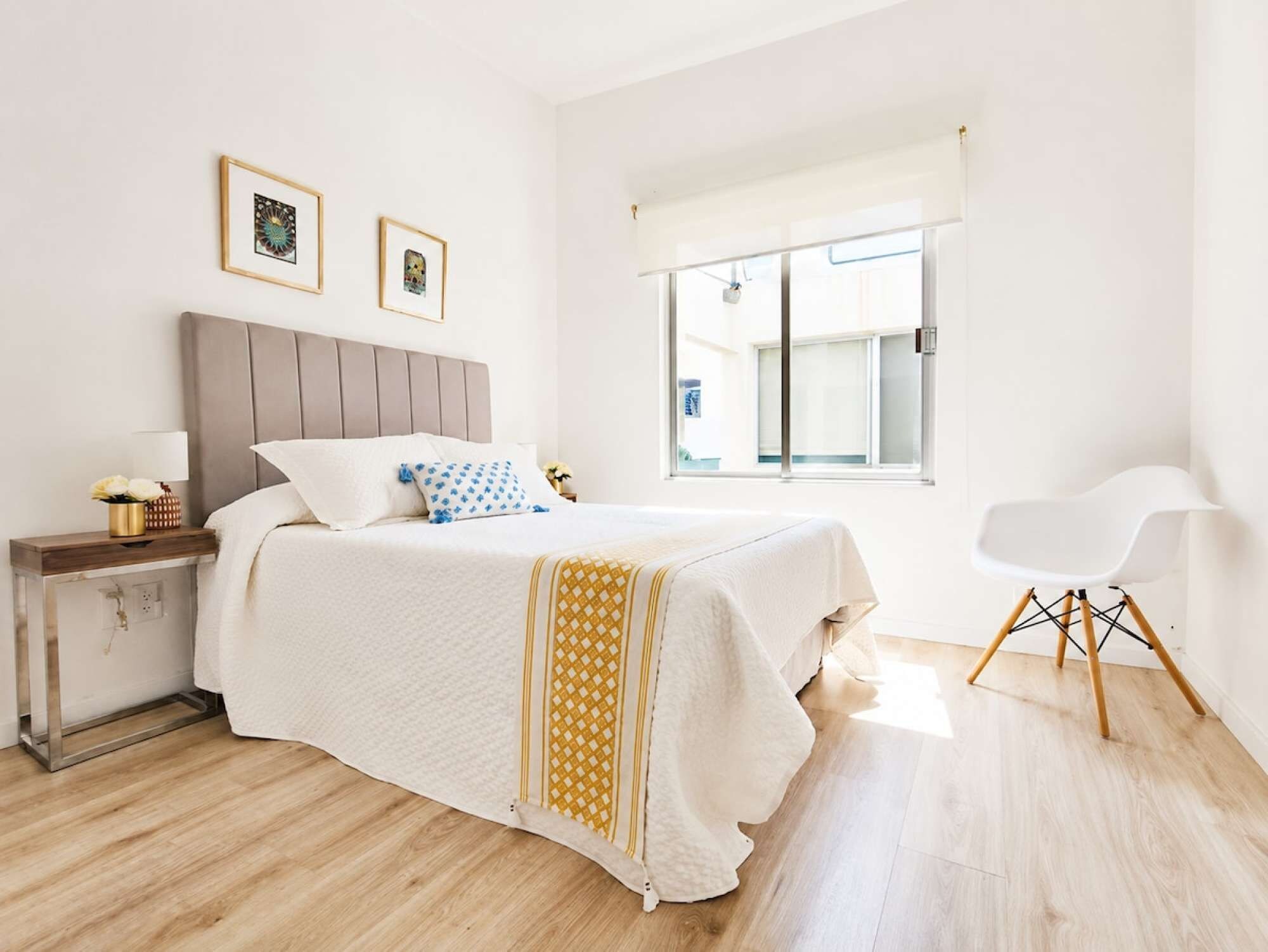 A bedroom with a bed, bedside table, lamp, chair, window, and artwork on the wall.