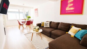Una sala moderna con un sofá marrón, un televisor, una zona de comedor con sillas rojas y un cartel con una frase motivadora.
