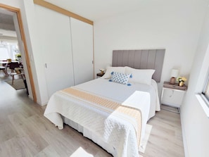 A bedroom with a bed, bedside tables, a nightstand, a window, and a door leading to another room.