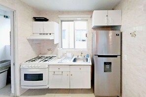 A compact kitchen with white cabinets, a stainless steel refrigerator, a gas stove, and a window with blinds.