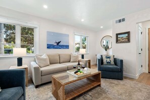 A living room with a beige sofa, a blue armchair, a glass coffee table with snacks, and artwork on the walls.