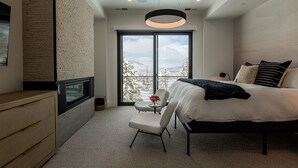 Ein Schlafzimmer mit einem großen Bett, einem Sessel, einem kleinen Tisch mit einer Vase und Blick auf schneebedeckte Berge durch ein großes Fenster.