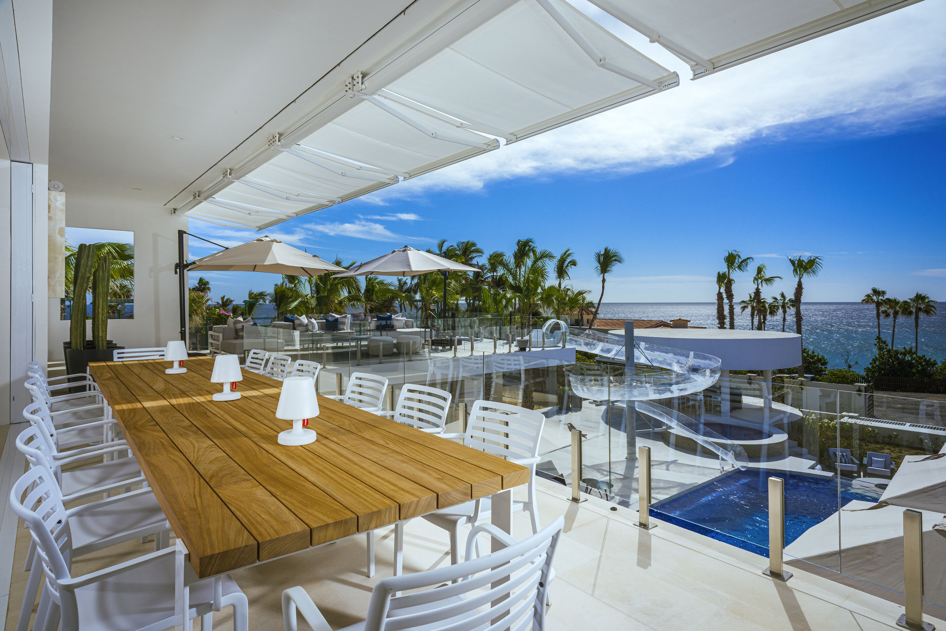 Een houten tafel met witte stoelen en parasols, met uitzicht op een zwembad en palmbomen.