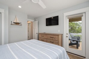 A bedroom with a bed, a dresser, a television, and a door leading to an outdoor area with chairs. - Shelly Shores - Hot tub, community pool & tennis courts, great views (Nags Head)