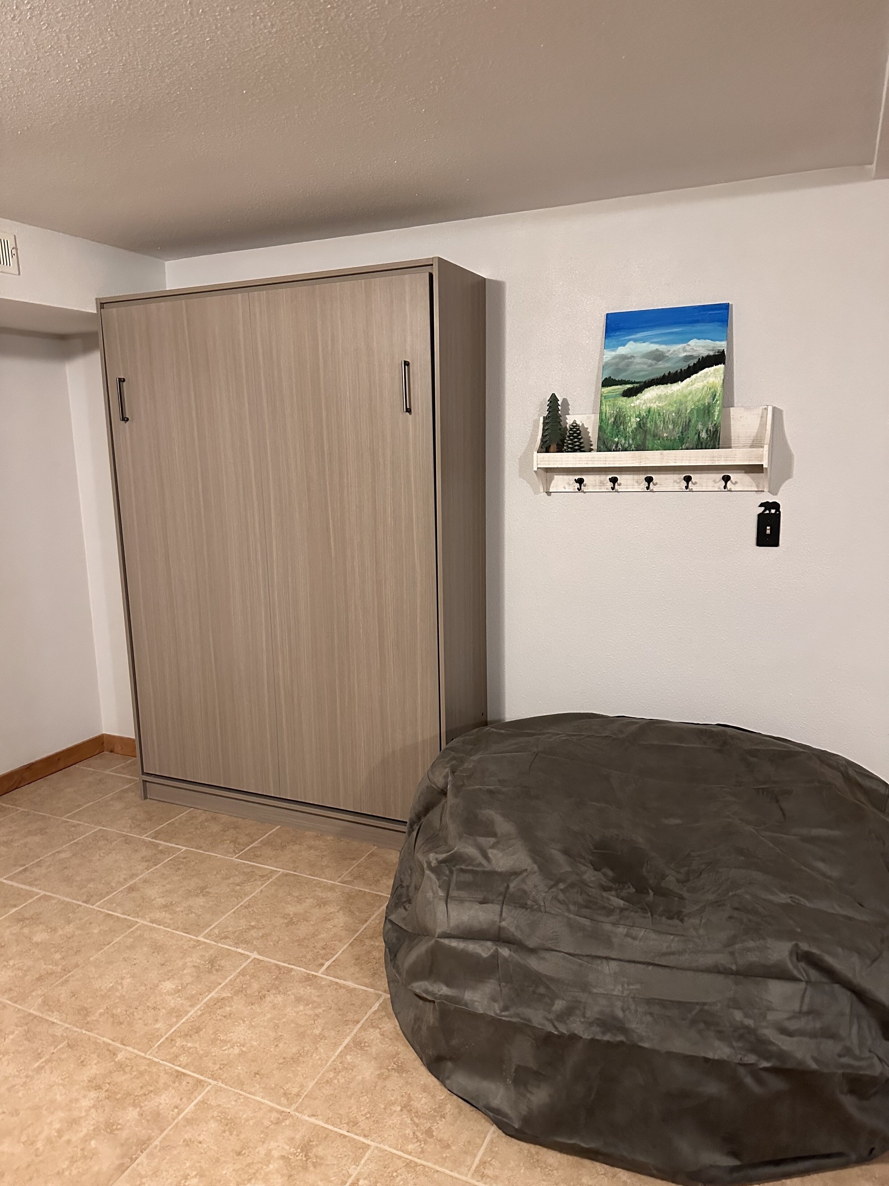 A room with a wardrobe, a bean bag, a wall-mounted shelf with a painting, and a small black object on the wall.