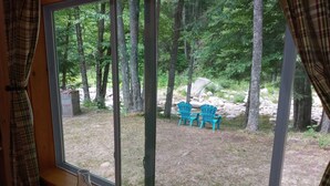 Une fenêtre donnant sur une zone boisée, deux chaises Adirondack bleues et une table en bois rustique avec une lampe.