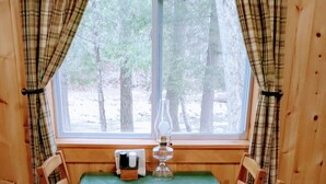 L’intérieur d’un chalet en bois, avec des rideaux à carreaux, une table recouverte d’une nappe verte, deux sets de table, une bougie et une fenêtre donnant sur les arbres.
