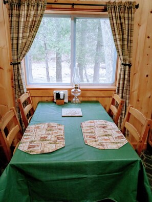 Interno di una baita in legno con tende a quadri, un tavolo con tovaglia verde, due sottopiatti, una candela e una finestra che si affaccia su alberi.