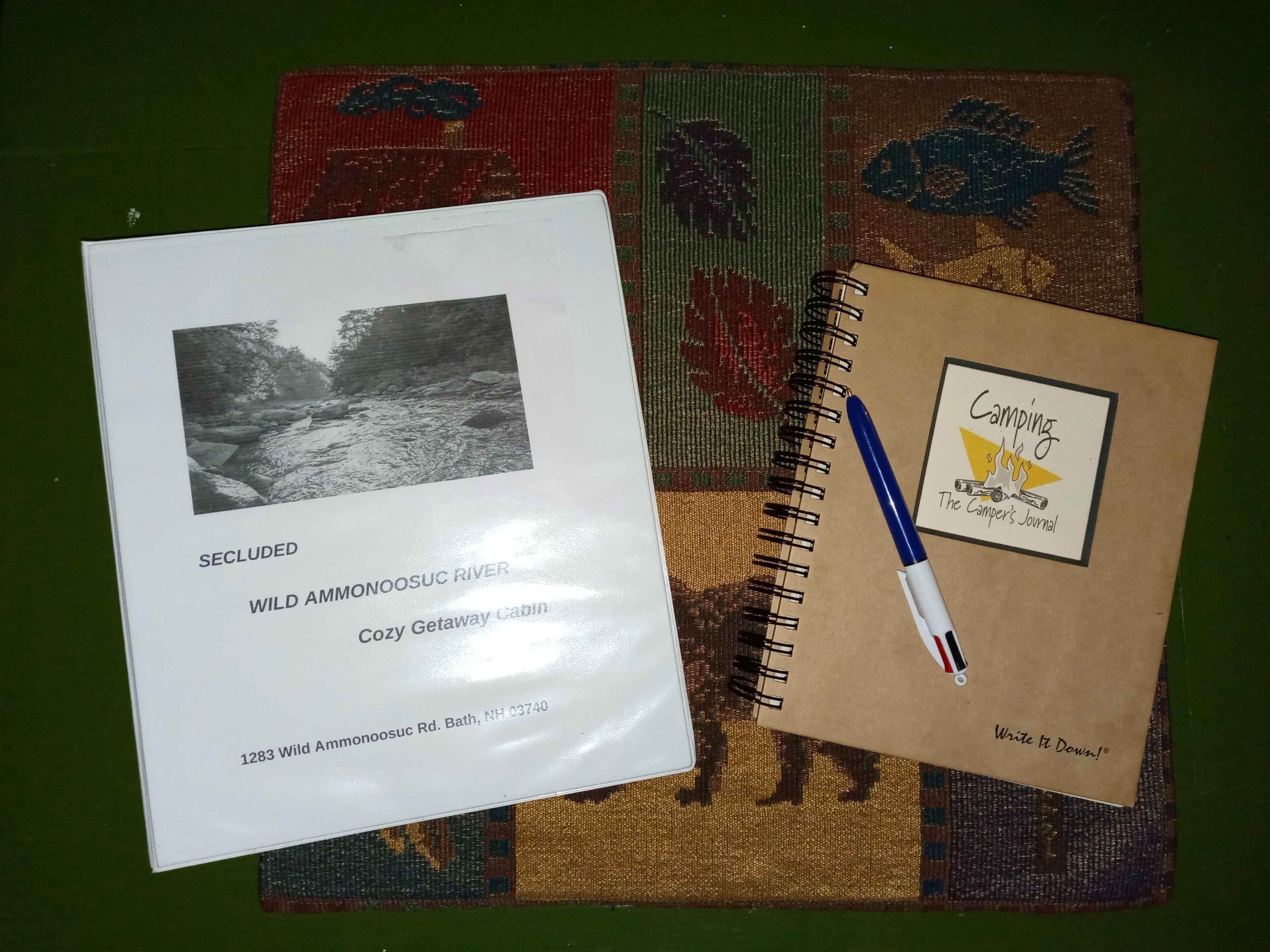 A booklet titled "SECLUDED WILD AMMONOOSUC RIVER COZY GETAWAY CABIN 1283 Wild Ammonoosuc Rd. Bath, NH 03740" and a notebook with a "Camping The Camper's Journal" logo.