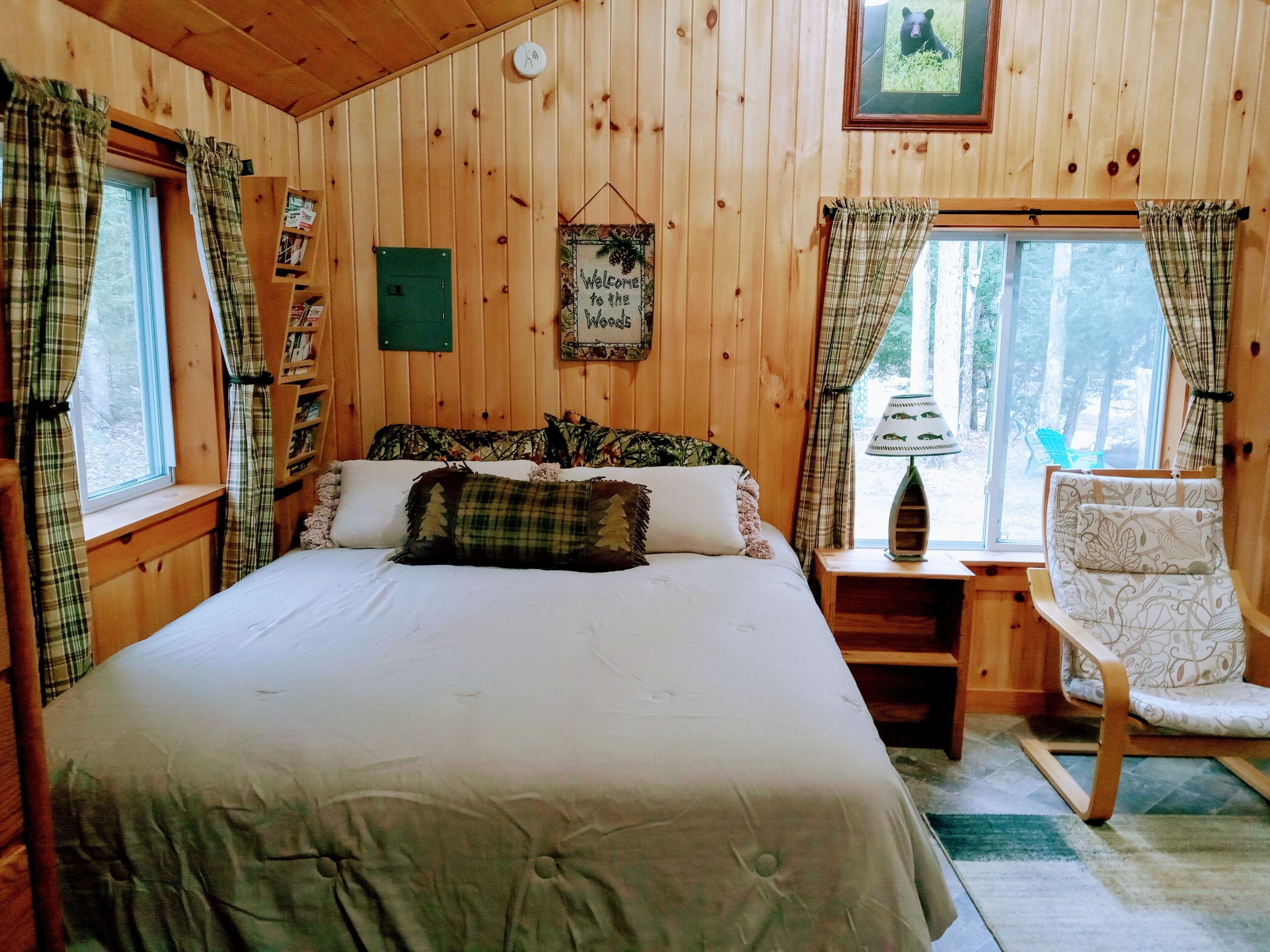 A wooden cabin bedroom with a bed, a chair, a nightstand, a window with curtains, a welcome sign, and a framed picture on the wall.