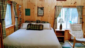 A wooden cabin bedroom with a bed, a chair, a nightstand, a window with curtains, a welcome sign, and a framed picture on the wall.