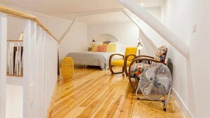 A cozy attic bedroom with a wooden floor, a bed, a chair, a fan, and a staircase.