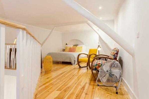 A cozy attic bedroom with a wooden floor, a bed, a chair, a fan, and a staircase.
