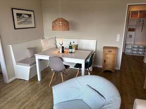 Ein Essbereich mit einem weißen Tisch, zwei Stühlen, einer Bank, einer Pendelleuchte und einem kleinen Schrank.