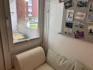 Une pièce dotée d’une fenêtre donnant sur un immeuble en briques, d’un petit balcon avec une table, et d’un mur décoré de diverses photographies et de notes.