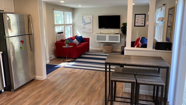 A modern kitchen with a stainless steel refrigerator, a red sofa, a flat-screen TV, and a dining area with a wooden table and benches. - Bright & Cozy, Yarmouth Cape Top Beaches! (Yarmouth)