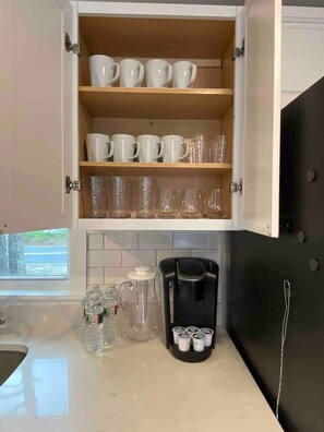 Kitchen features a white cabinet with mugs and clear glass cups, a countertop with a coffee maker and water dispenser, and a window with an outside view.
