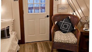 Une chambre mansardée chaleureuse, avec une porte blanche, un fauteuil en osier agrémenté d’un coussin à motifs et un ventilateur posé sur une table en bois.