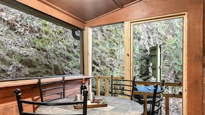 A wooden cabin with a stone-tiled table and chairs, a wooden deck, and a view of a forested hillside. - Cozy Family Cabin, Hot Tub, Game Room, 2 King Beds Near Dollywood, Pet Friendly (Pigeon Forge)