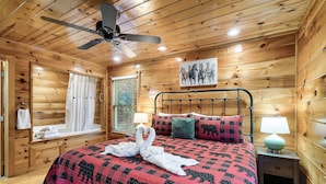 A wooden cabin-style room with a bed, a ceiling fan, a window with curtains, a wall-mounted picture, and a nightstand with a lamp. - Cozy Family Cabin, Hot Tub, Game Room, 2 King Beds Near Dollywood, Pet Friendly (Pigeon Forge)