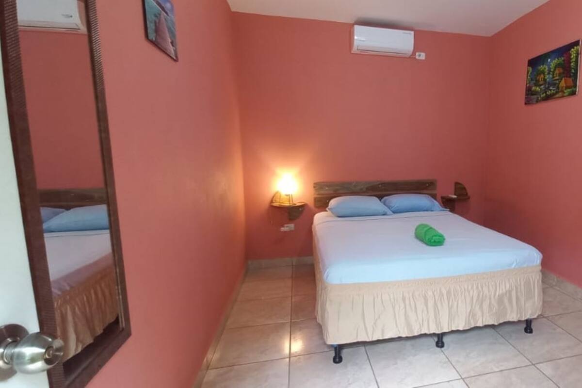 Quarto com cama, mesa de cabeceira, ar condicionado fixado na parede, porta e luminária na parede.