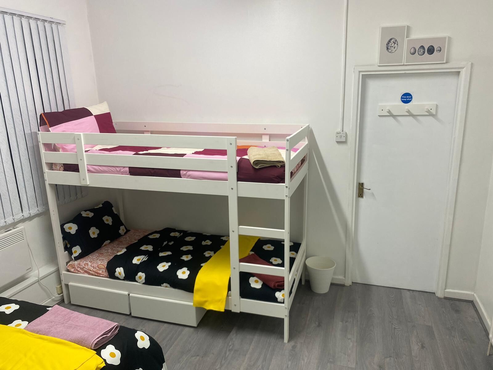 A bunk bed room with a door, a window with blinds, and a wall-mounted light switch.