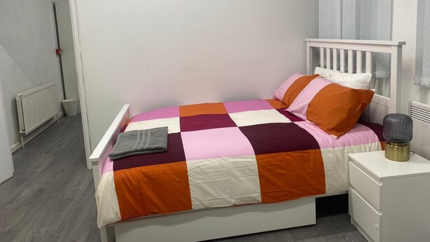 Quarto individual com uma cama de solteiro, cabeceira branca, mesa de cabeceira com luminária e radiador na parede.