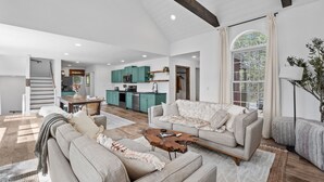 A spacious living area with a large sofa, a coffee table, and a dining table. There is a kitchen in the background with teal cabinets and a staircase leading upstairs. - kids secret room, Canoes/Kayaks, fire pit, ev charger. (Center Harbor)
